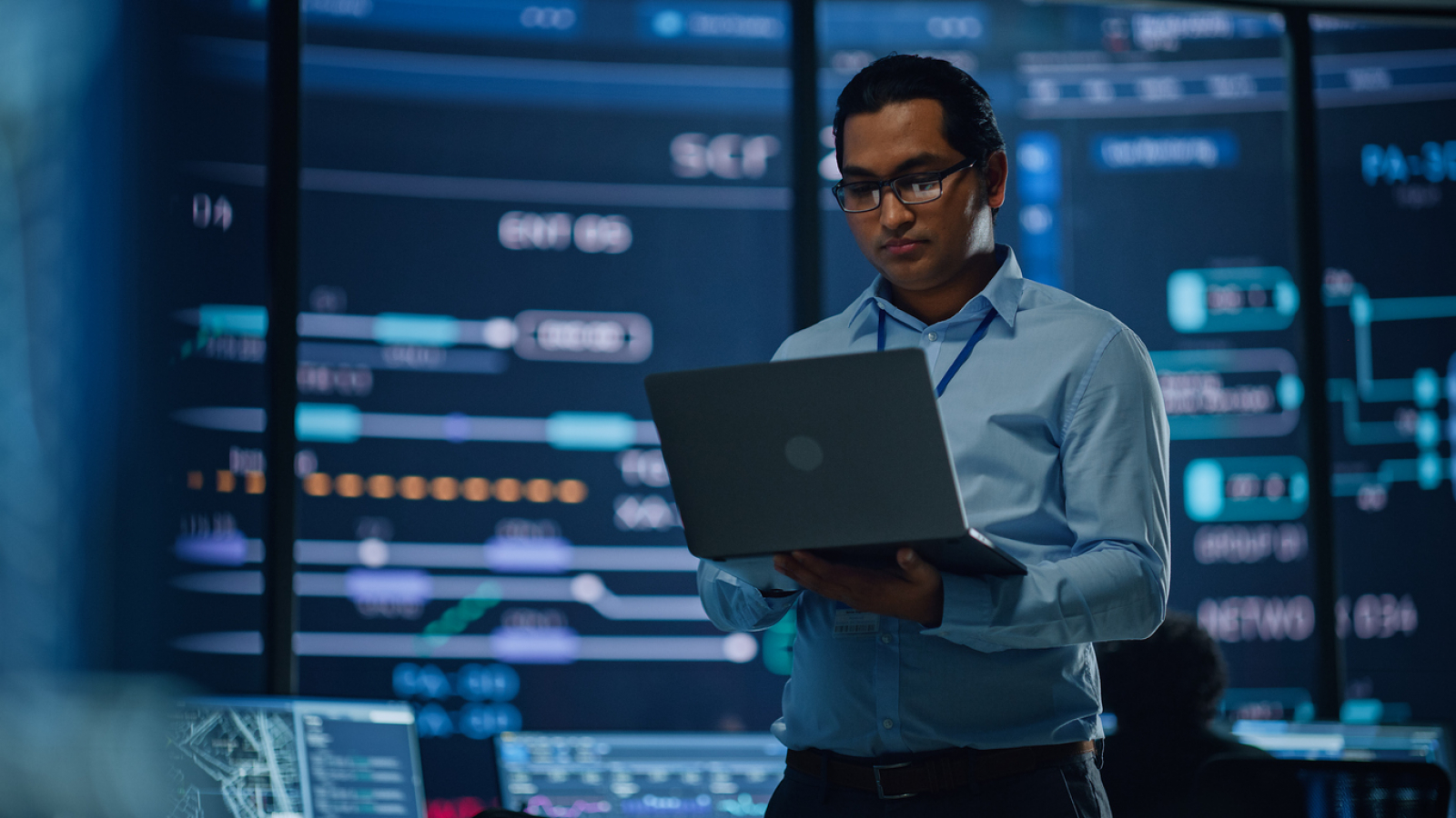 Young Multiethnic Male Employee Uses Laptop Computer in System Control Monitoring Center. In the Background His Coworkers at Their Workspaces with Many Displays Showing Technical Data.