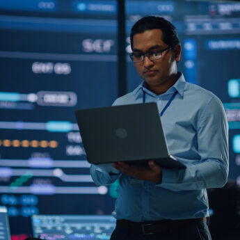 Young Multiethnic Male Employee Uses Laptop Computer in System Control Monitoring Center. In the Background His Coworkers at Their Workspaces with Many Displays Showing Technical Data.