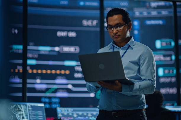 Young Multiethnic Male Employee Uses Laptop Computer in System Control Monitoring Center. In the Background His Coworkers at Their Workspaces with Many Displays Showing Technical Data.