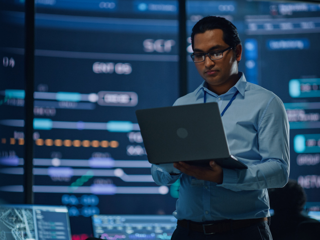 Young Multiethnic Male Employee Uses Laptop Computer in System Control Monitoring Center. In the Background His Coworkers at Their Workspaces with Many Displays Showing Technical Data.
