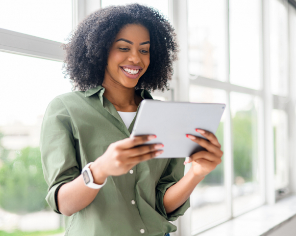 An African-American woman with a beaming smile uses a tablet, engaging in a video call or browsing digital content, standing by a window that floods the room with natural light.