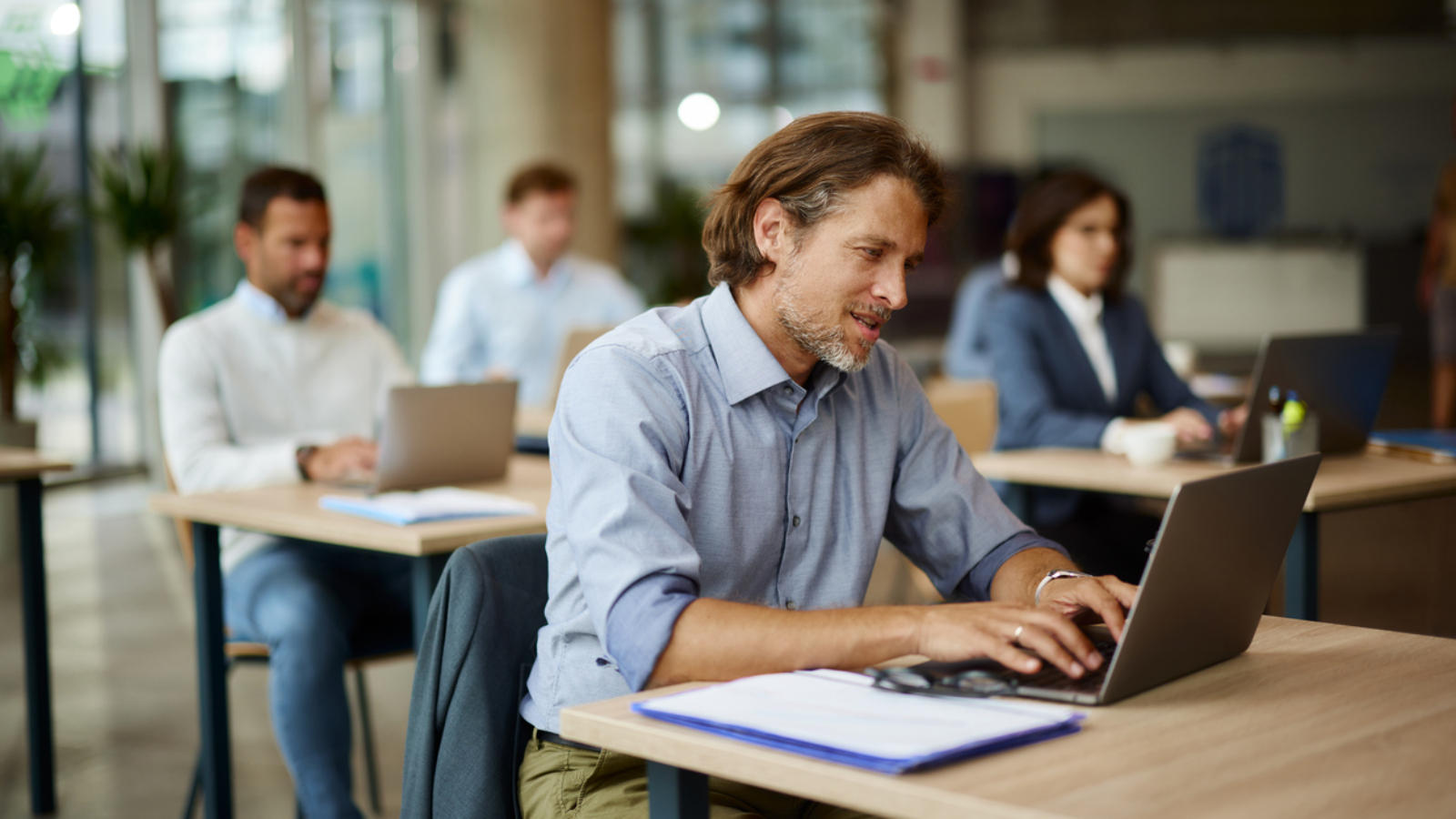 Mid adult businessman typing an e-mail on a computer in the office. His colleagues are in the background.