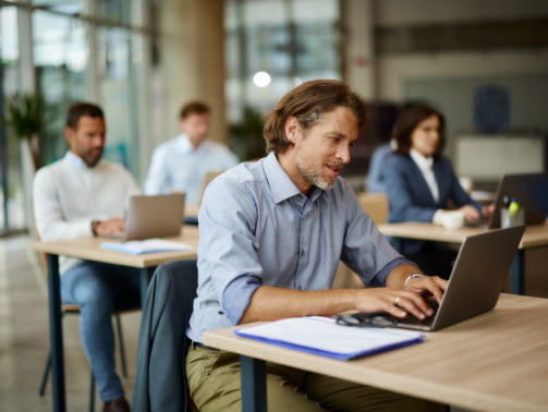 Mid adult businessman typing an e-mail on a computer in the office. His colleagues are in the background.