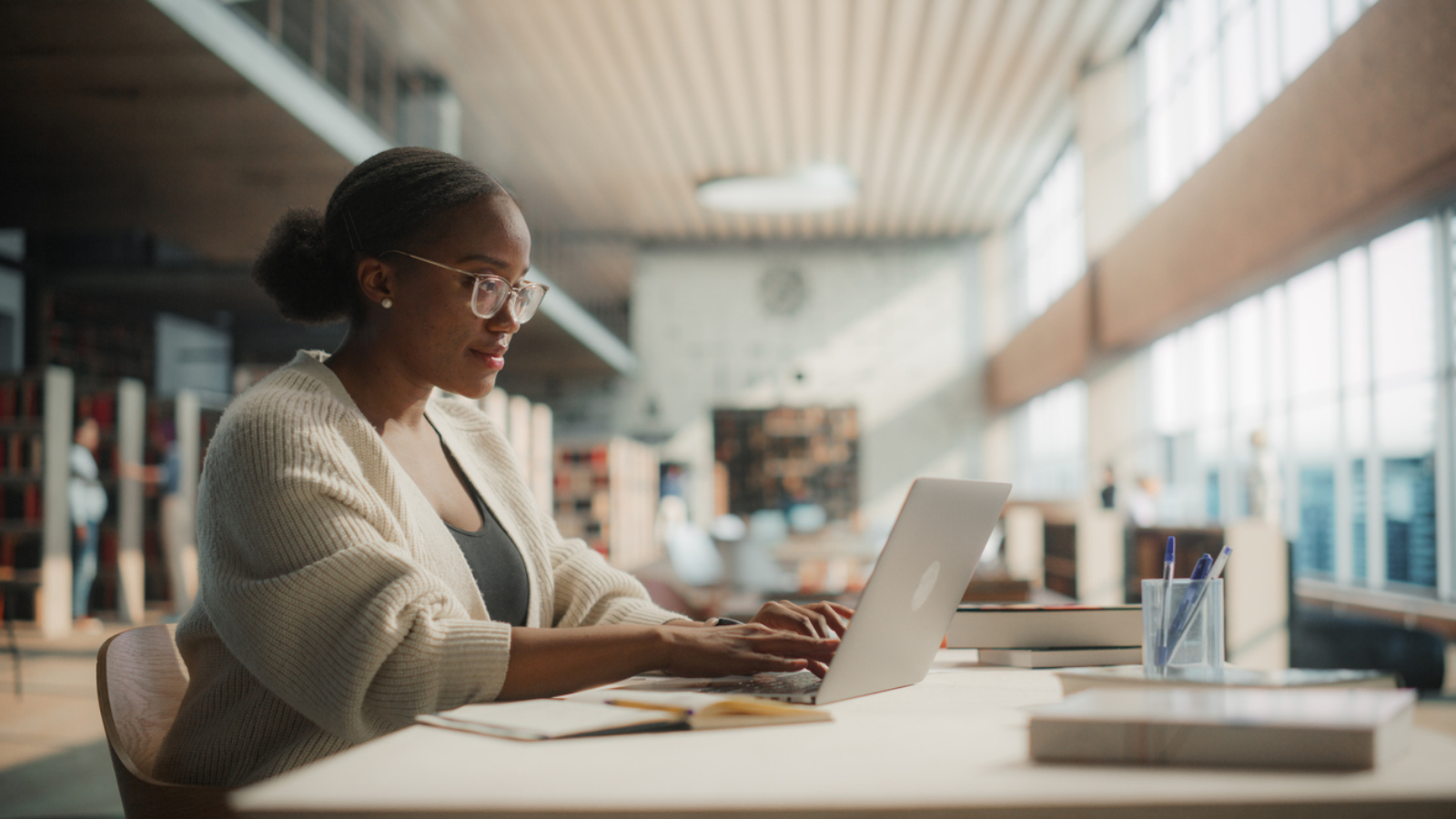 Dedicated African American Female Student Engaged in Online Learning at a Modern Library. Young Woman Using Laptop for Research, Surrounded by Books and Academic Environment.