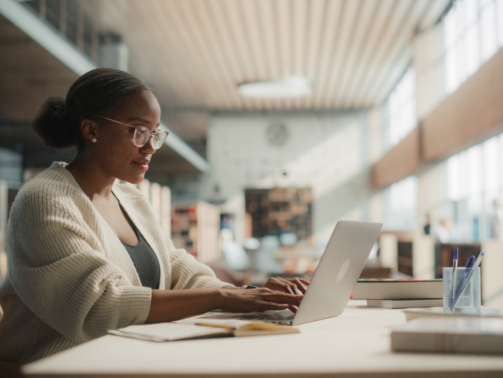 Dedicated African American Female Student Engaged in Online Learning at a Modern Library. Young Woman Using Laptop for Research, Surrounded by Books and Academic Environment.
