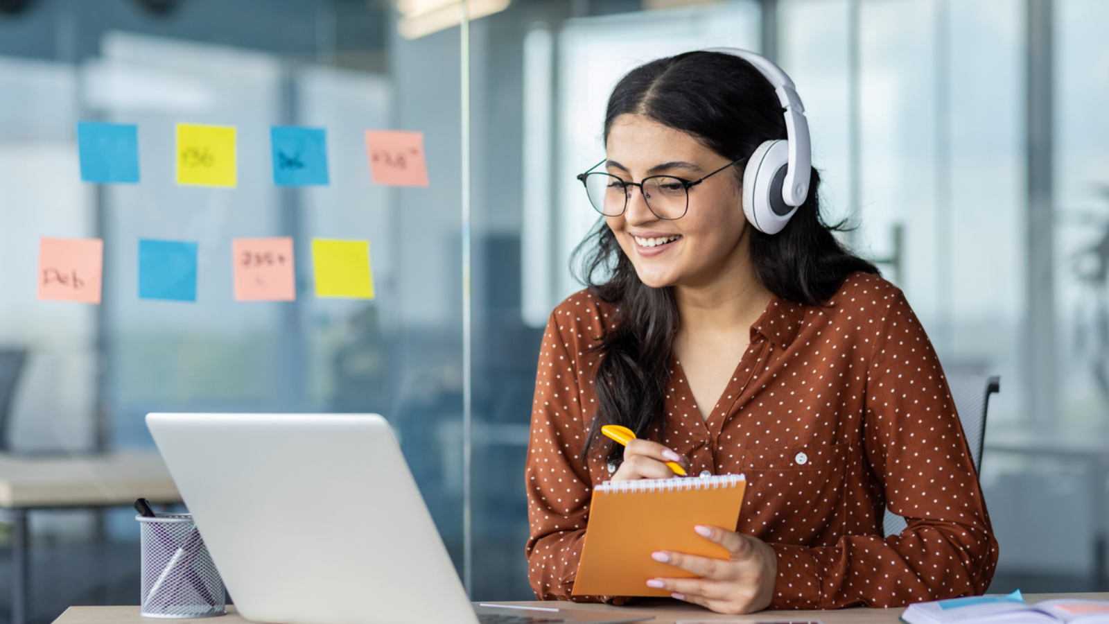 Woman watching online video course, training conference inside office at workplace. Office worker in headphones using laptop for remote meeting, writing data in notebook.
