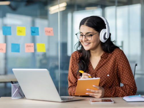 Woman watching online video course, training conference inside office at workplace. Office worker in headphones using laptop for remote meeting, writing data in notebook.