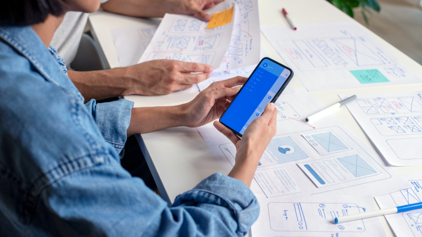 UX/UI designer testing prototype on a phone, discussing and brainstorming on wireframes for a website and mobile app prototype, surrounded by sketches of user flow and design tools, in the concept of website and mobile application design concept.