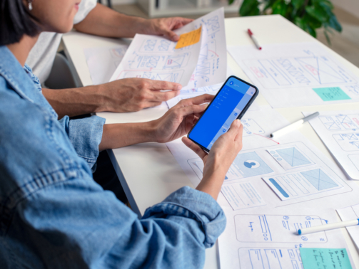 UX/UI designer testing prototype on a phone, discussing and brainstorming on wireframes for a website and mobile app prototype, surrounded by sketches of user flow and design tools, in the concept of website and mobile application design concept.