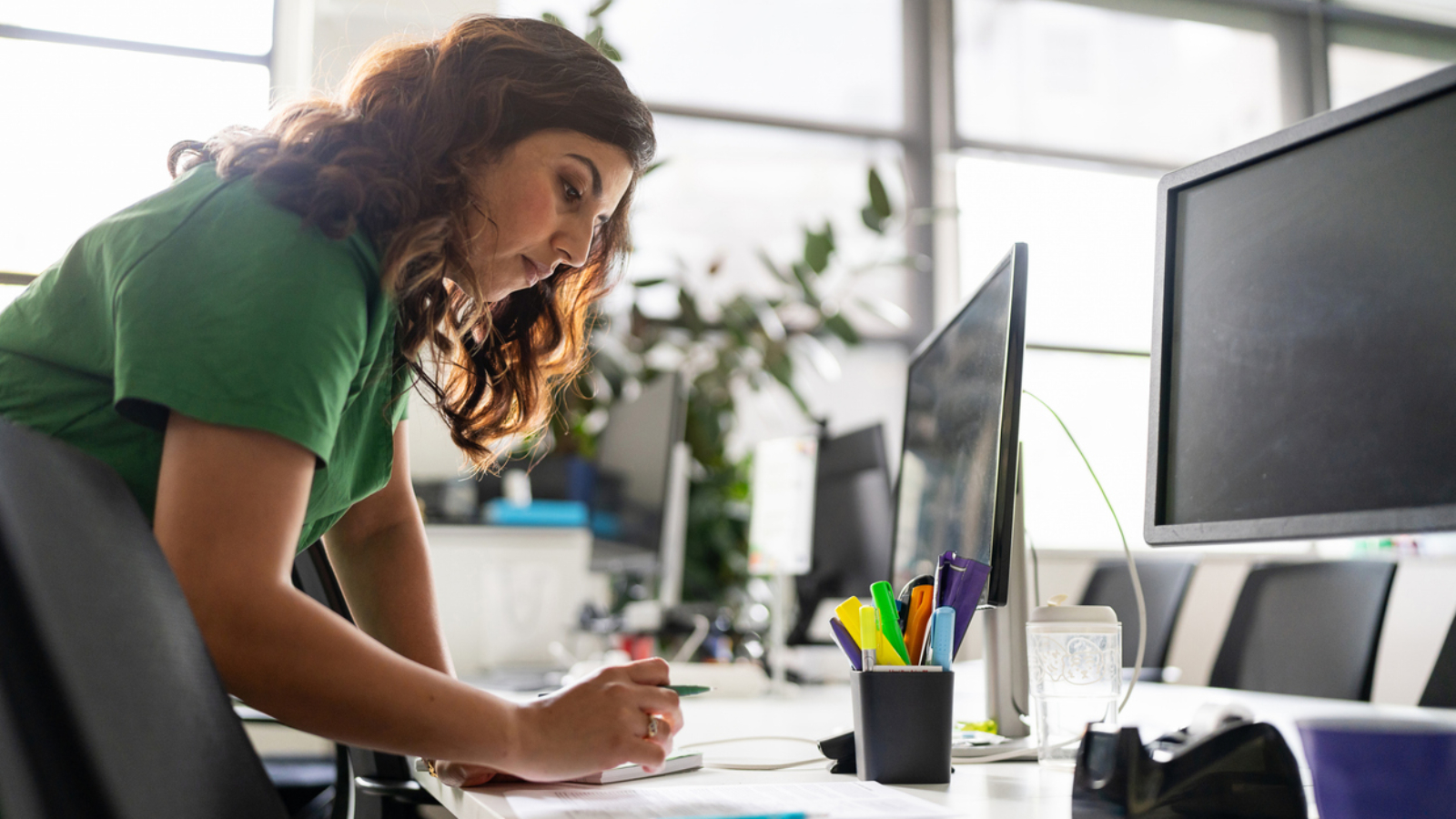 A focused professional woman working on documents at an office desk, showcasing dedication and productivity in a modern workspace environment, surrounded by office tools and natural light.
