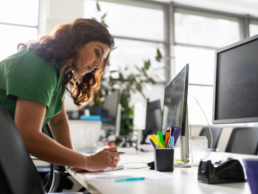 A focused professional woman working on documents at an office desk, showcasing dedication and productivity in a modern workspace environment, surrounded by office tools and natural light.