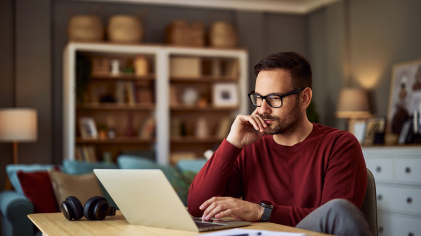 A busy, focused young adult male freelancer sitting at a desk and resting his head on his hand while working on a project on his laptop.