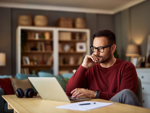 A busy, focused young adult male freelancer sitting at a desk and resting his head on his hand while working on a project on his laptop.