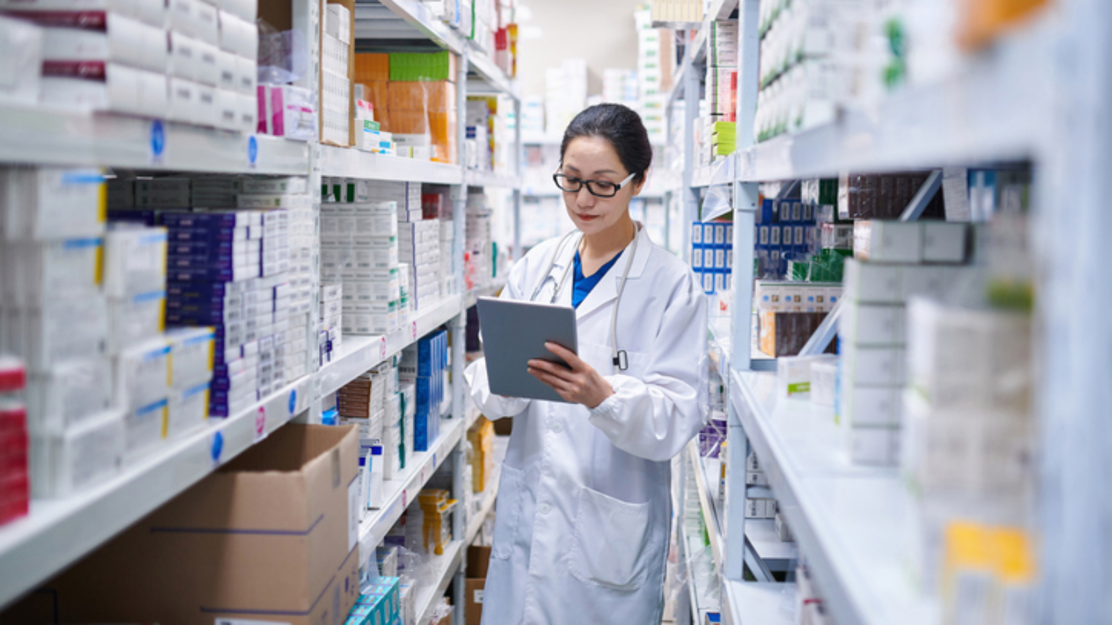 Side view of one asia female pharmacist using digital tablet while taking inventory at pharmacy warehouse.
