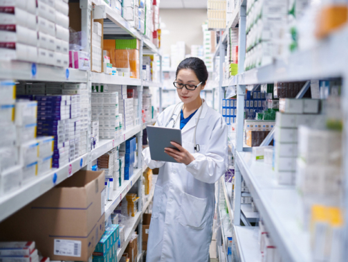 Side view of one asia female pharmacist using digital tablet while taking inventory at pharmacy warehouse.