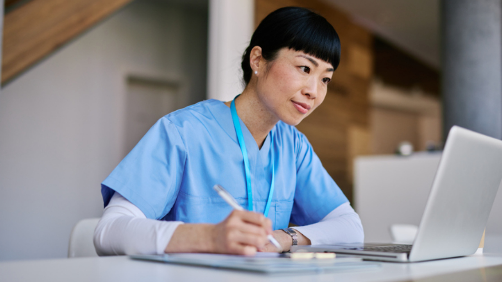 Mid adult woman nurse wearing blue scrubs, taking notes and focused on her laptop while working in a hospital environment, contributing to healthcare and patient care efforts