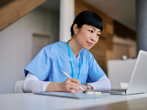 Mid adult woman nurse wearing blue scrubs, taking notes and focused on her laptop while working in a hospital environment, contributing to healthcare and patient care efforts