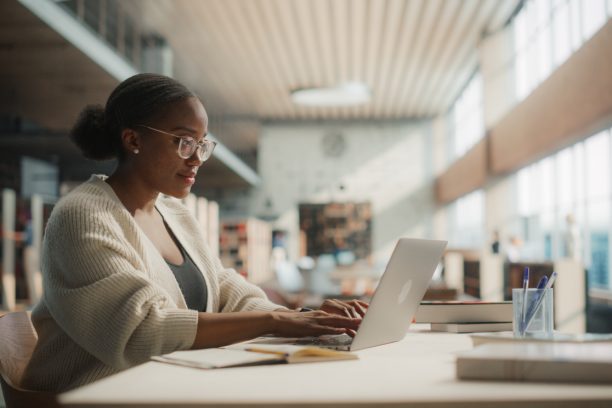 Dedicated African American Female Student Engaged in Online Learning at a Modern Library. Young Woman Using Laptop for Research, Surrounded by Books and Academic Environment.