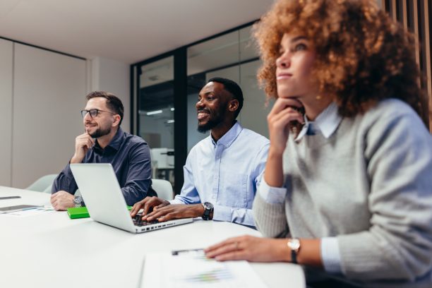 Group of businesspeople engaged in a corporate meeting, listening attentively to a presentation while a man takes notes on his laptop, fostering collaboration and teamwork