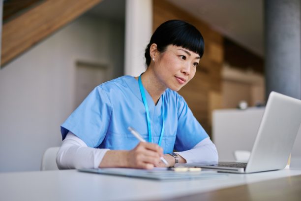 Mid adult woman nurse wearing blue scrubs, taking notes and focused on her laptop while working in a hospital environment, contributing to healthcare and patient care efforts